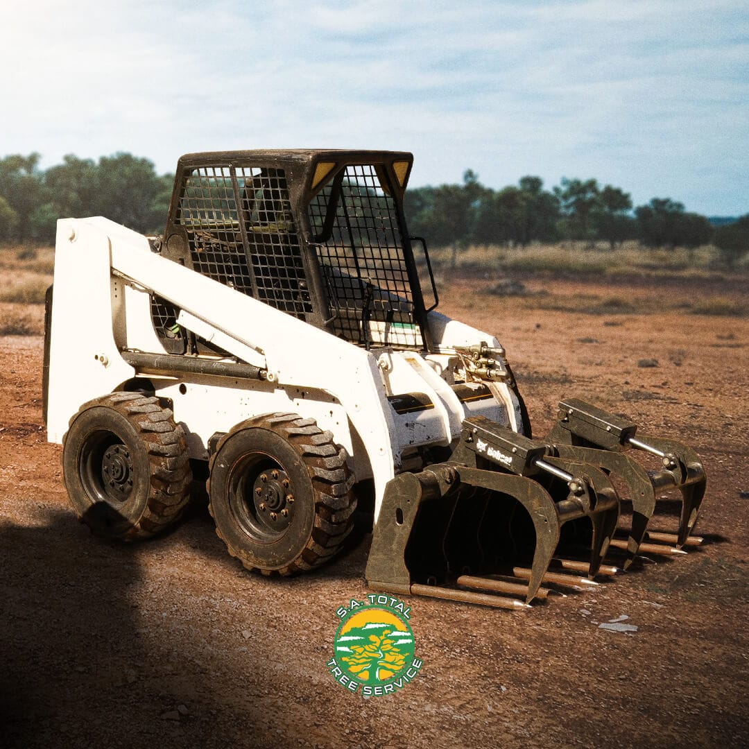 A white skid steer loader with a grapple attachment is parked on a dirt surface. The logo of "A Total Tree Care Company" is visible in front of the loader. Trees are seen in the background.